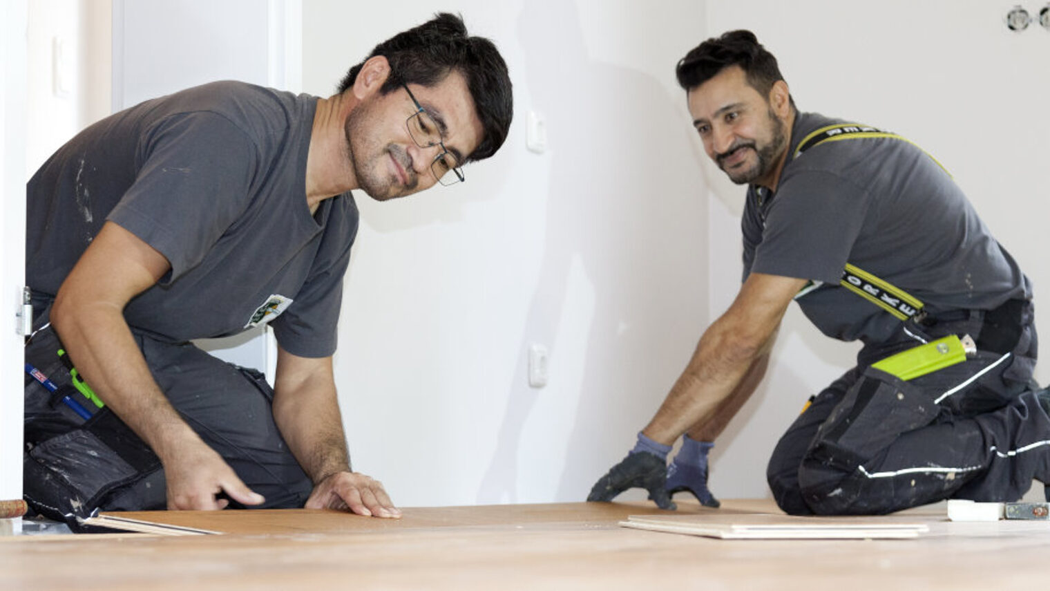 Zwei Männer in Arbeitskleidung verlegen knieend in einem Zimmer mit weißen Wänden einen Parkettboden.