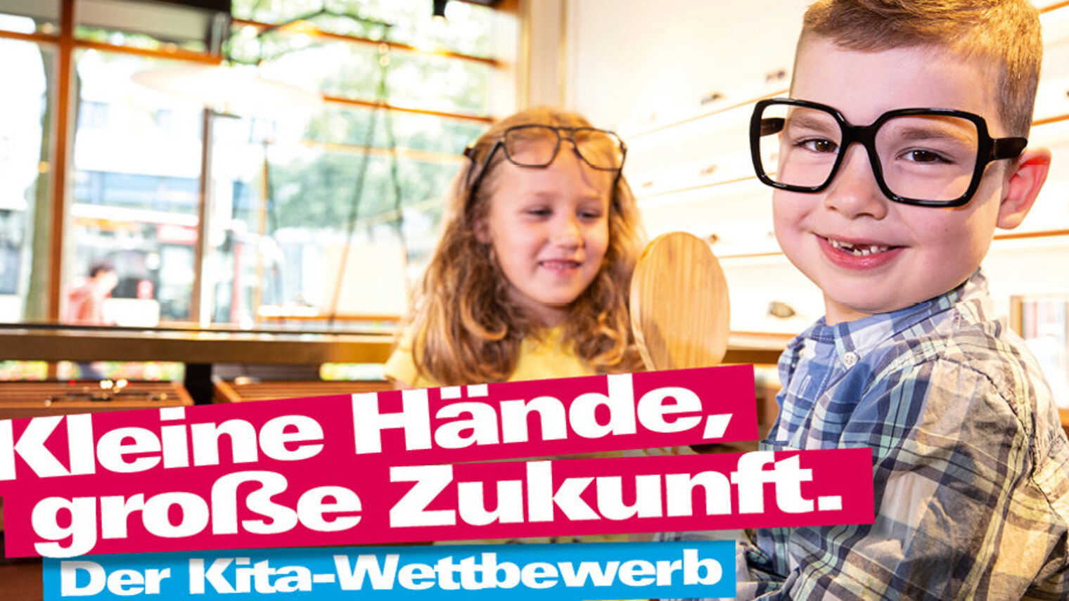 Zwei Kinder probieren in einem Augenoptiker-Geschäft Brillen, Schriftzug "Kleine Hände, große Zukunft. Der Kita-Wettbewerb"