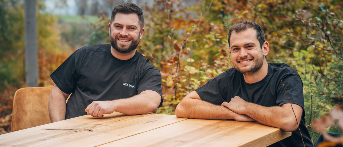 Zwei junge Männer in schwarzen T-Shirts sitzen im Freien an einem Holztisch und lächeln in die Kamera.