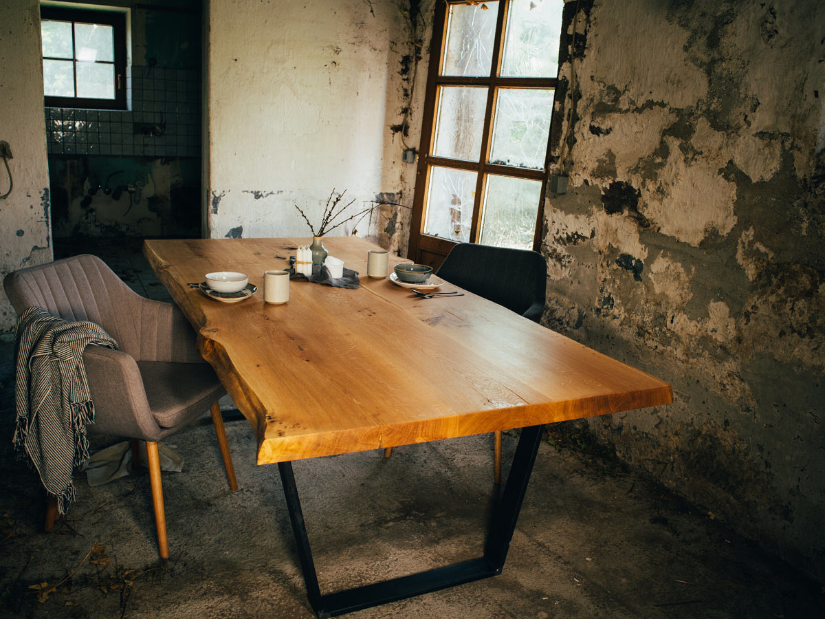 Holztisch, Stuhl und etwas Geschirr stehen in einem Raum eines alten Hauses mit kaputten Fenstern und bröckelndem Putz. 
