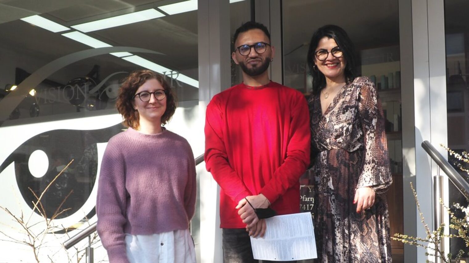Zwei Frauen und ein junger Mann stehen nebeneinander vor Eingang eines Friseursalons mit Glasfront und lächeln in die Kamera
