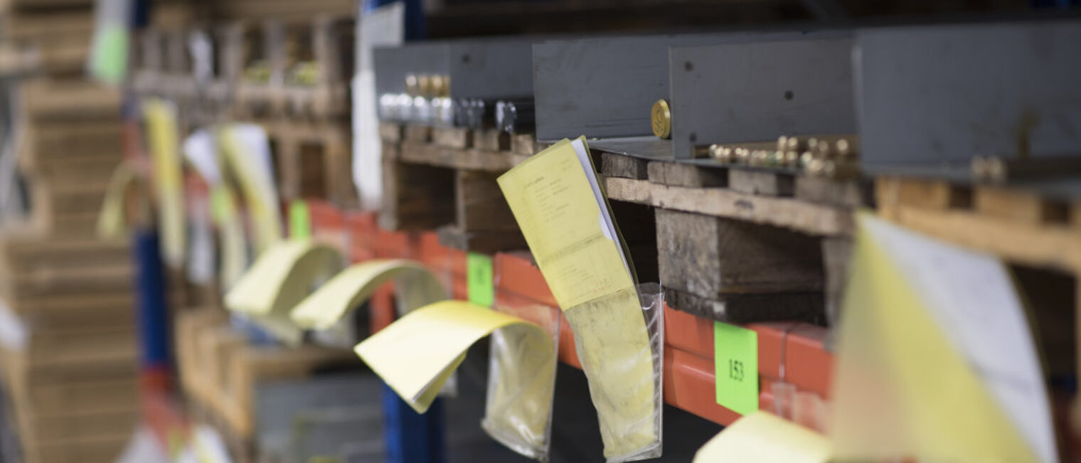 In einem Lager für Material sieht man viele Fächer auf Paletten, in denen Metallstäbe liegen. Davor gelbe Zettel und Nummern.