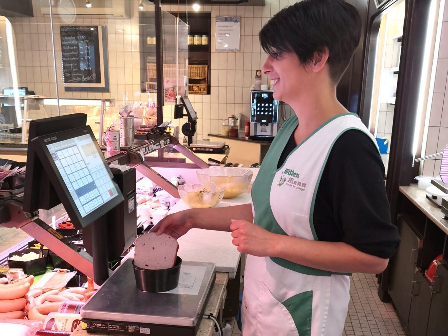 Junge Frau mit kurzen dunklen Haaren legt eine Scheibe Leberkäse in eine schwarze runde Box auf einer Waage in der Metzgerei.