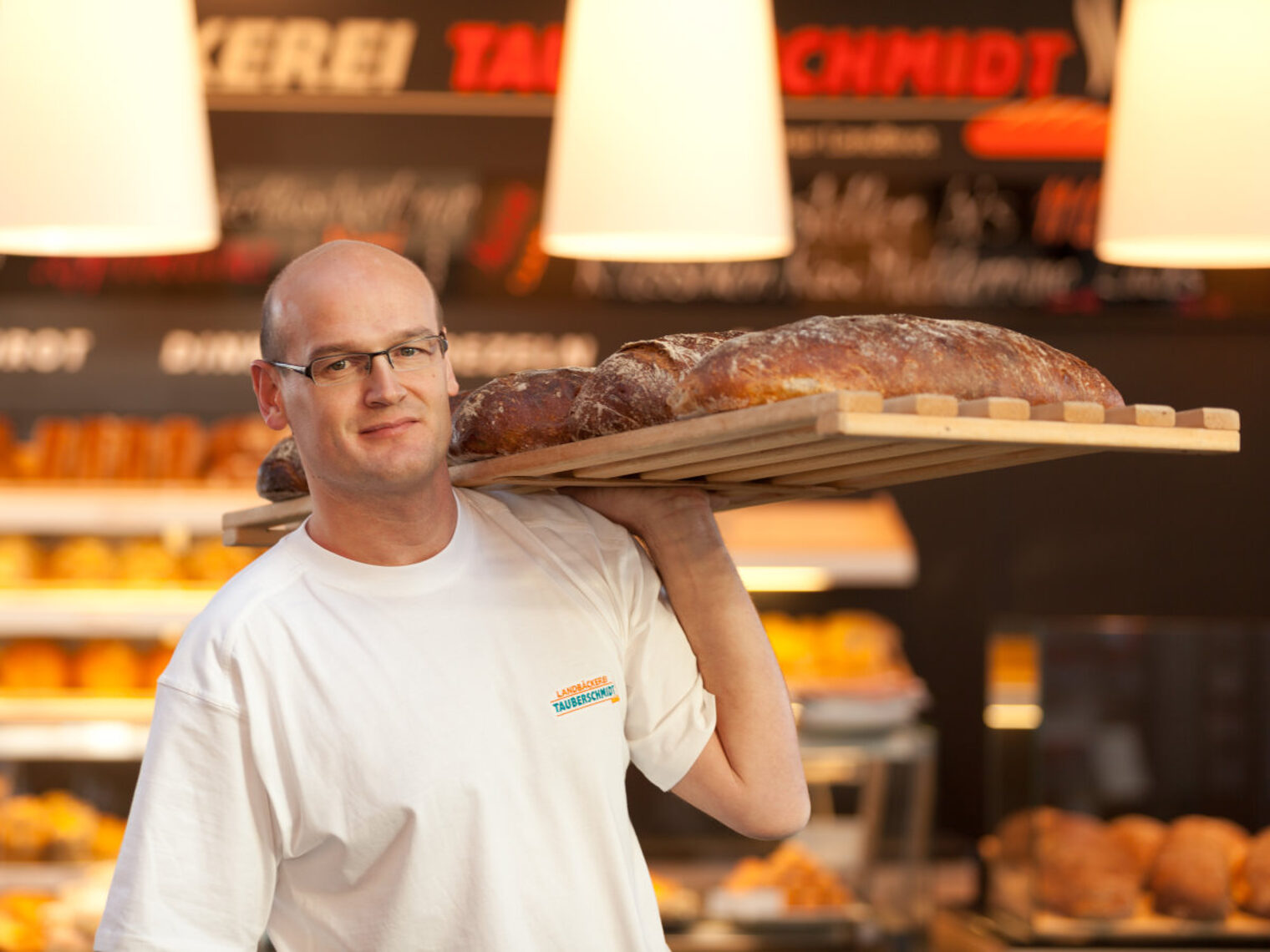 Mann in weißem T-Shirt steht in einer Bäckerei und trägt ein großes Brett mit Broten gestützt von einer Hand auf der Schulter