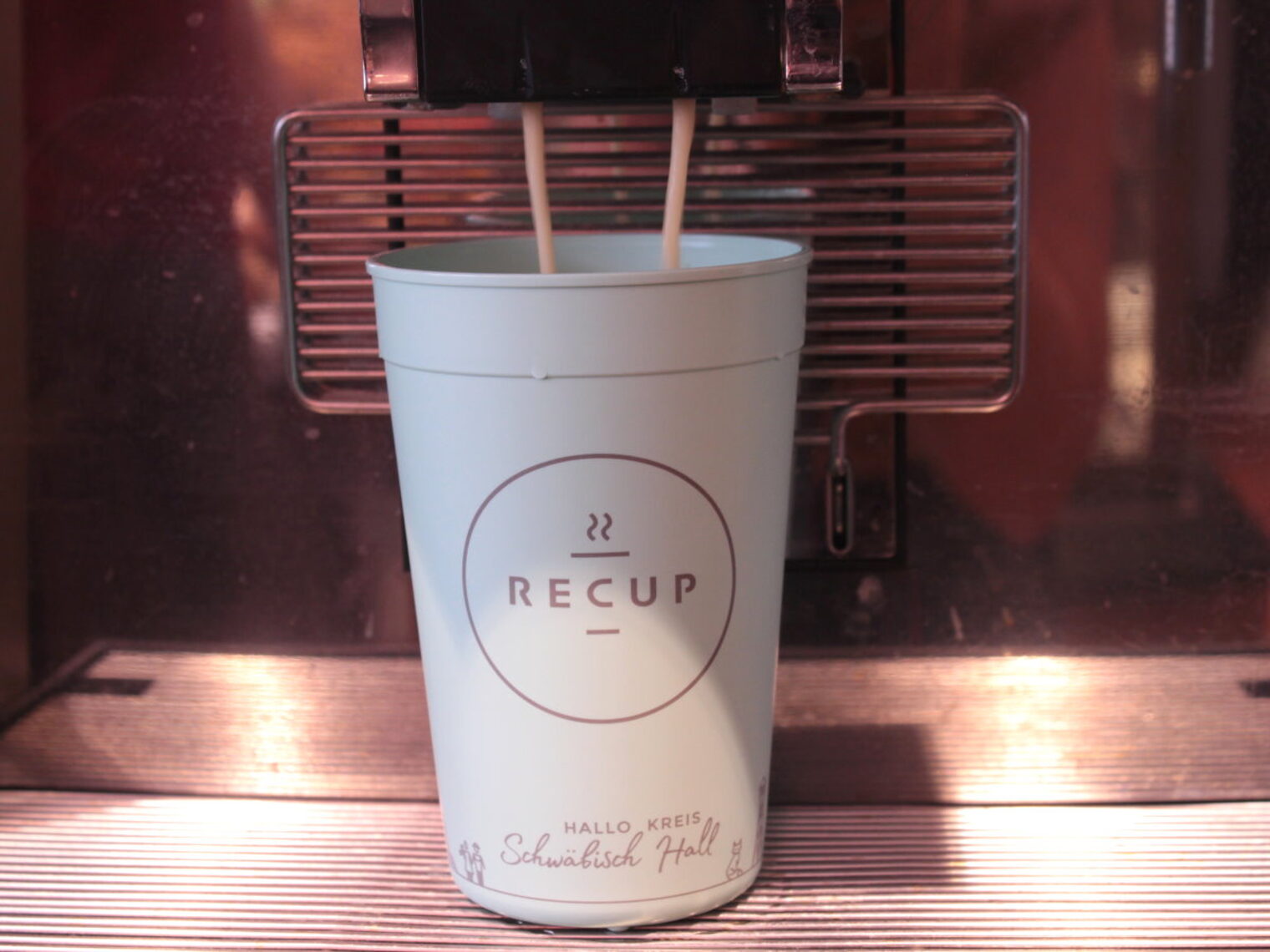 Ein hellblauer Mehrweg-Becher mit Logo Recup und Schriftzug Schwäbisch Hall wird in einer Kaffemaschine mit Kaffee befüllt. 