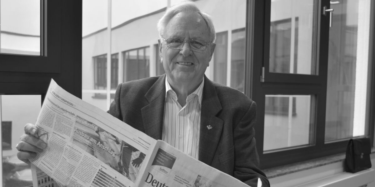 Älterer Mann mit Brille sitzt vor Fenster mit einer Zeitung in der Hand, dargestellt in schwarz-weiß.