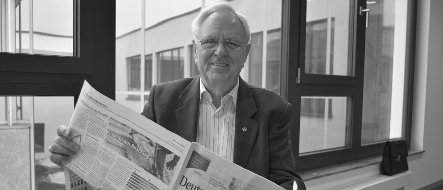 Älterer Mann mit Brille sitzt vor Fenster mit einer Zeitung in der Hand, dargestellt in schwarz-weiß.