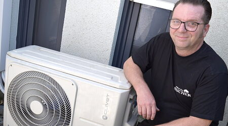 Mann mit Brille und schwarzem T-Shirt kniet neben dem Außengerät einer Klimanlage, auf die er den rechten Arm gestützt hat.