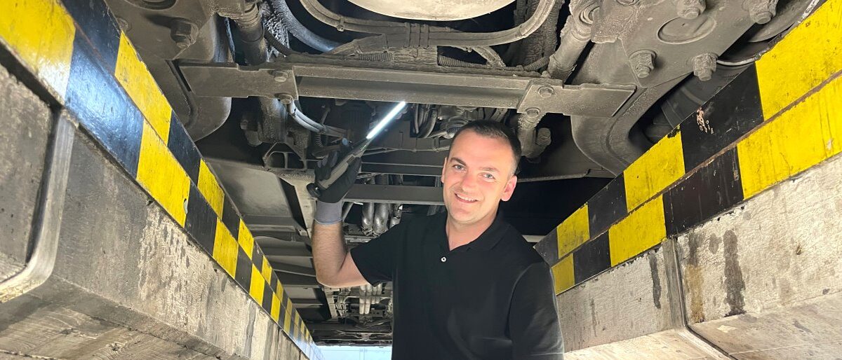 Kfz-Mechatroniker-Meister Mentor Musa prüft einem Bus. Junger Mann in schwarzem Polohemd steht in Grube einer Werkstatt unter einem Fahrzeug, Leuchte in der Hand, lächelt in Kamera.