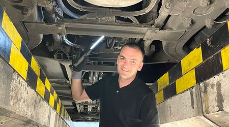 Junger Mann in schwarzem Polohemd steht in Grube einer Werkstatt unter einem Fahrzeug, Leuchte in der Hand, lächelt in Kamera.