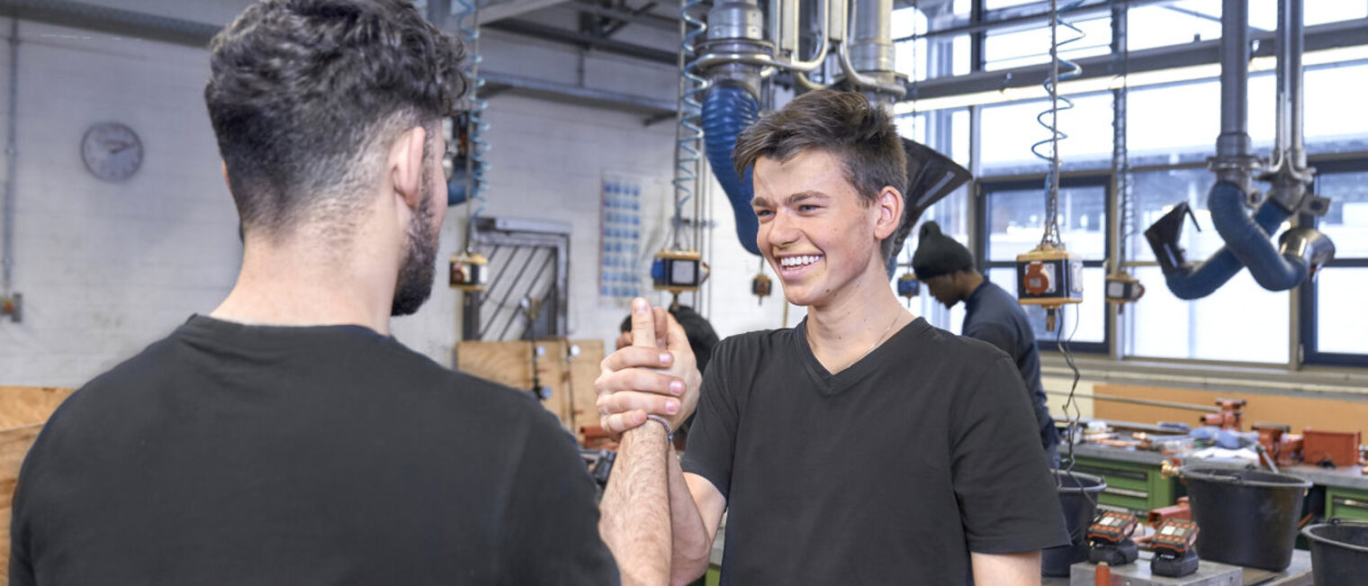 Zwei junge Männer in schwarzen T-Shirts begrüßen sich in einer Werkstatt lachend mit einem Handschlag.