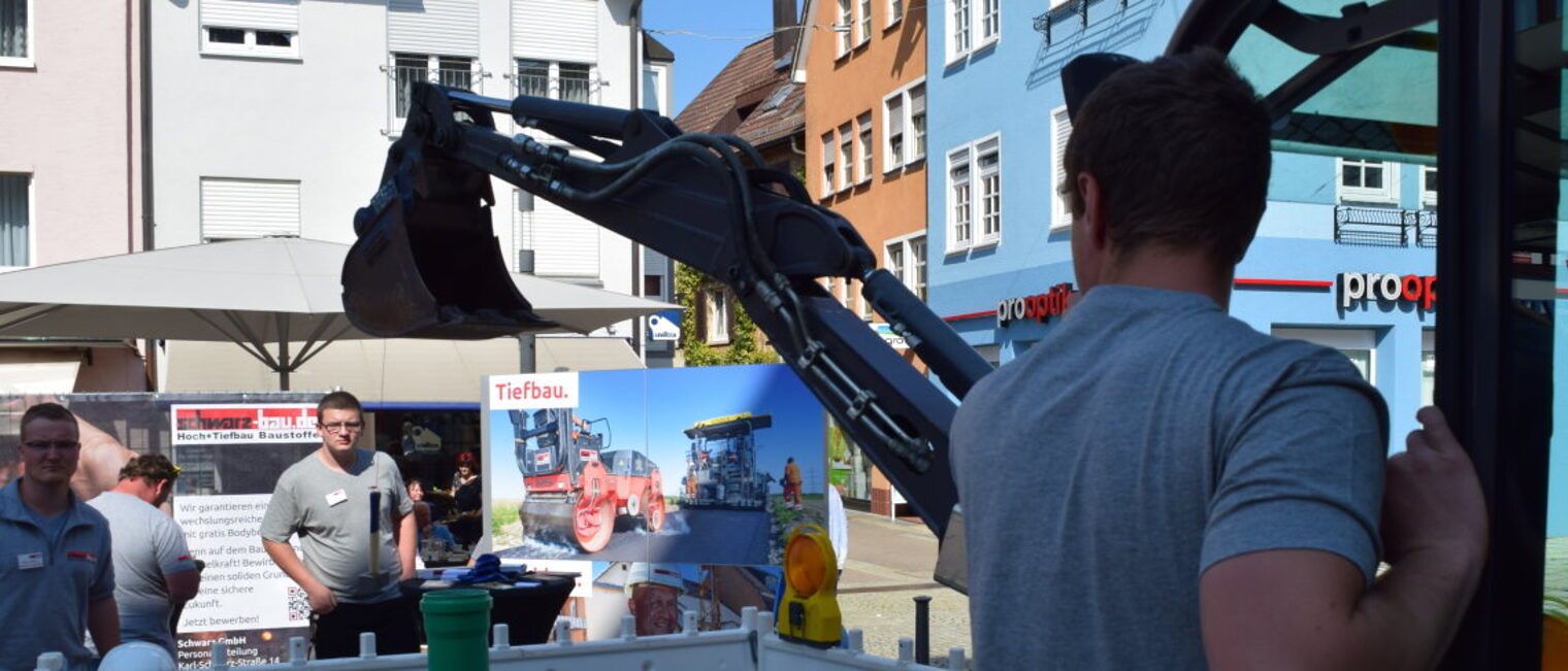 Beim Handwerkertag gibt es Einblicke in verschiedene Handwerksberufe . Ein Mann steht in einer Fußgängerzone an einem kleinen Bagger und erklärt interessierten Jugendlichen, wie er funktioniert.