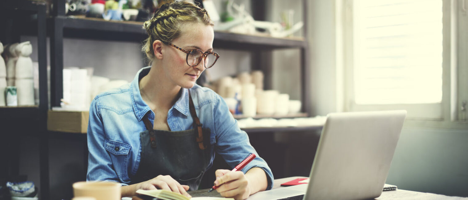 Eine junge Keramikerin sitzt mit einem Laptop am Schreibtisch und macht sich Notizen.
