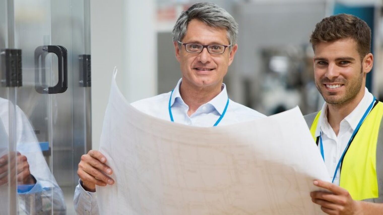 Zwei Männer halten gemeinsam einen Plan und lächeln in die Kamera.