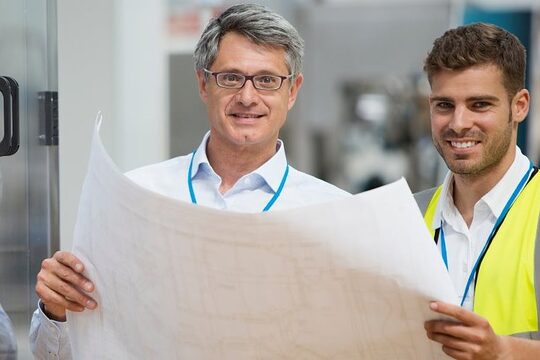 Zwei Männer halten gemeinsam einen Plan und lächeln in die Kamera.