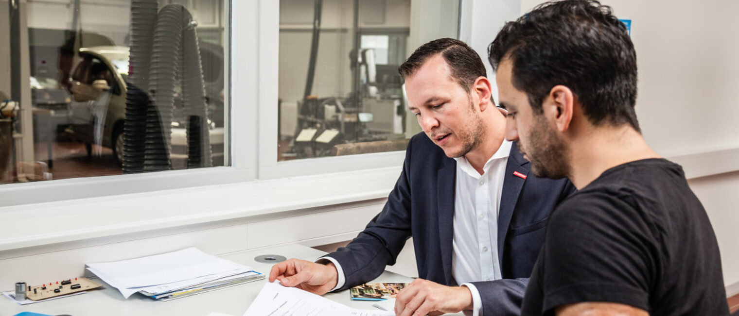 Für den Notfall vorbereitet - Tipps gibt es im Web-Seminar am 20. Juli. Zwei Männer sitzen im Büro einer Autowerkstatt. Sie unterhalten sich und schauen auf Unterlagen auf dem Tisch.