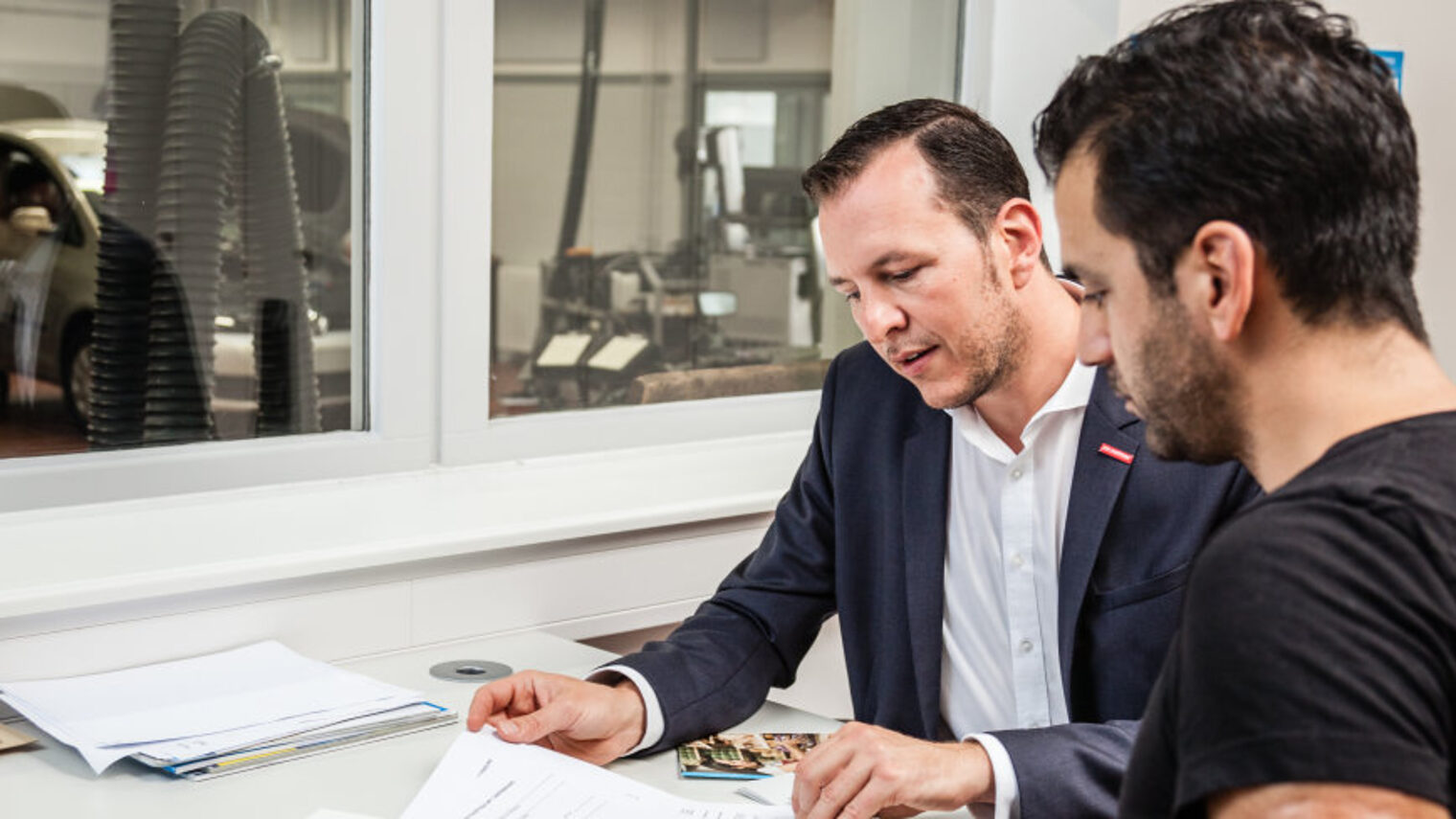 Zwei Männer sitzen im Büro einer Autowerkstatt. Sie unterhalten sich und schauen auf Unterlagen auf dem Tisch.