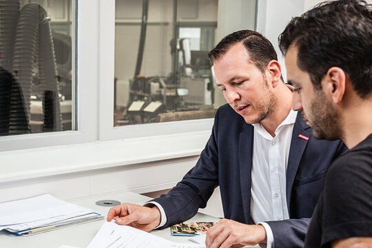 Zwei Männer sitzen im Büro einer Autowerkstatt. Sie unterhalten sich und schauen auf Unterlagen auf dem Tisch.