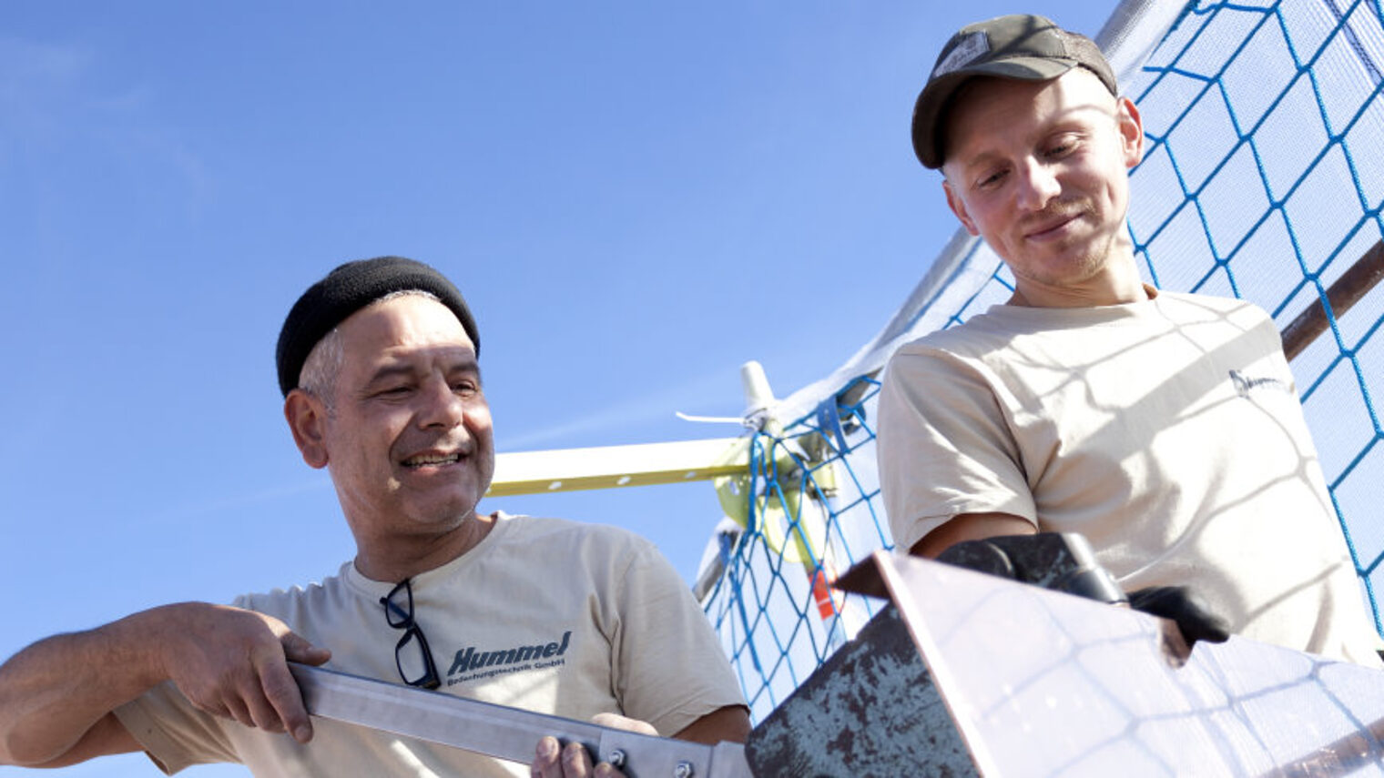 Zwei Männer stehen vor einem Schutznetz und bearbeiten ein Blech.