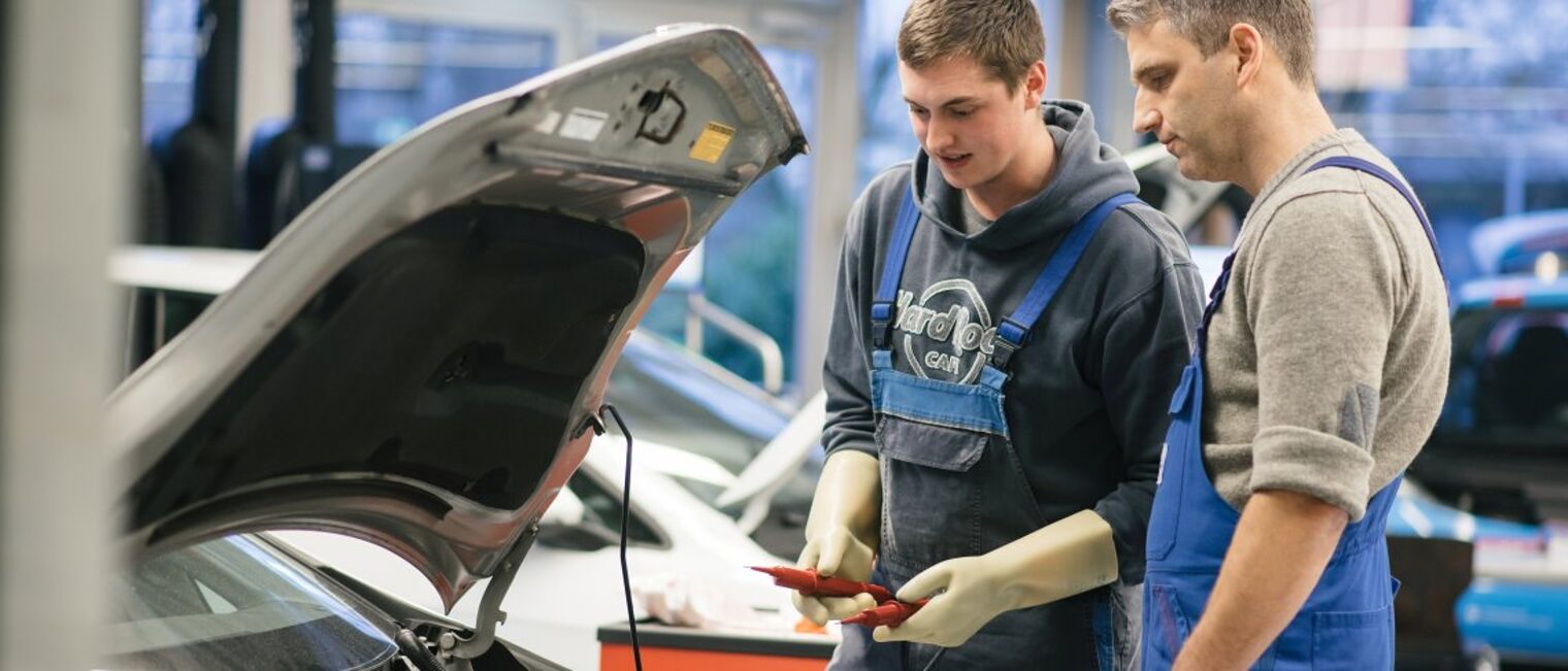 Ein älterer und ein jüngerer Mann in Latzhosen blicken in den Motorraum eines Autos. 