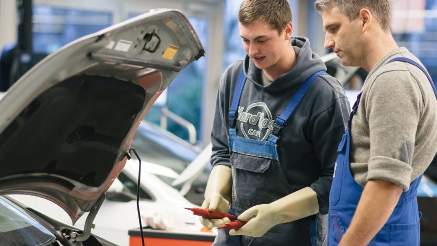 Ein älterer und ein jüngerer Mann in Latzhosen blicken in den Motorraum eines Autos. 