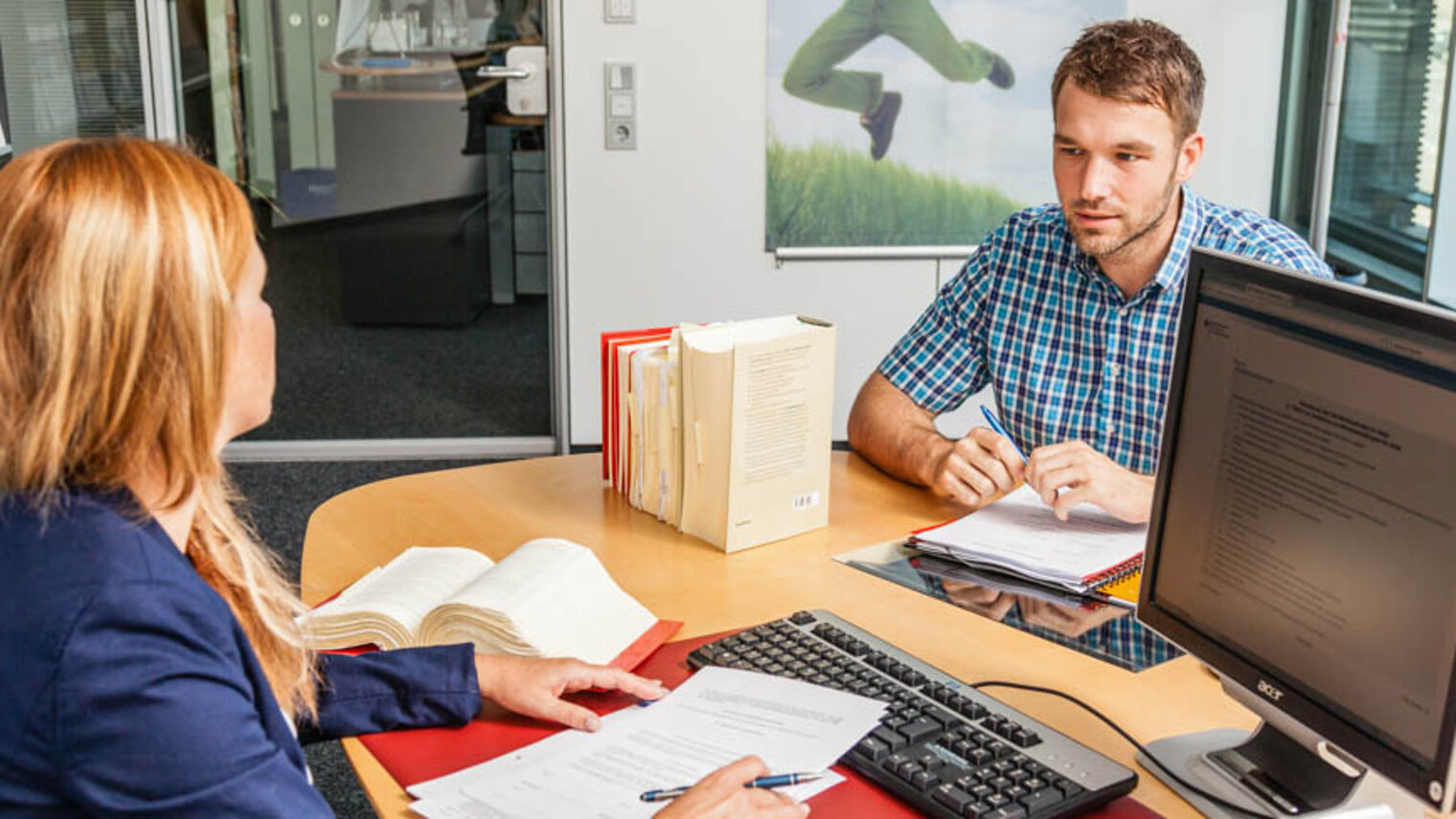 Ein Mann sitzt zur Beratung bei einer Frau am Tisch. Vor der Frau liegt ein Vertrag sowie Gesetzesbücher.