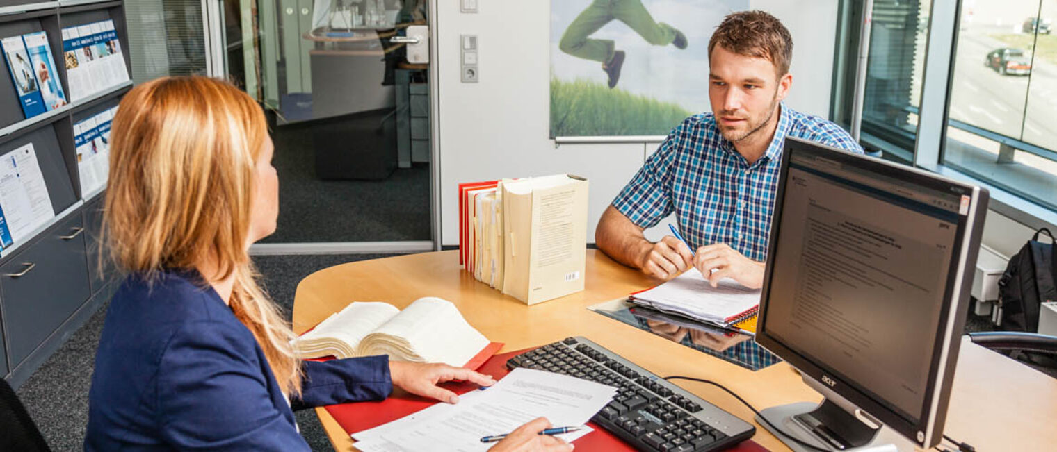 Ein Mann sitzt zur Beratung bei einer Frau am Tisch. Vor der Frau liegt ein Vertrag sowie Gesetzesbücher.