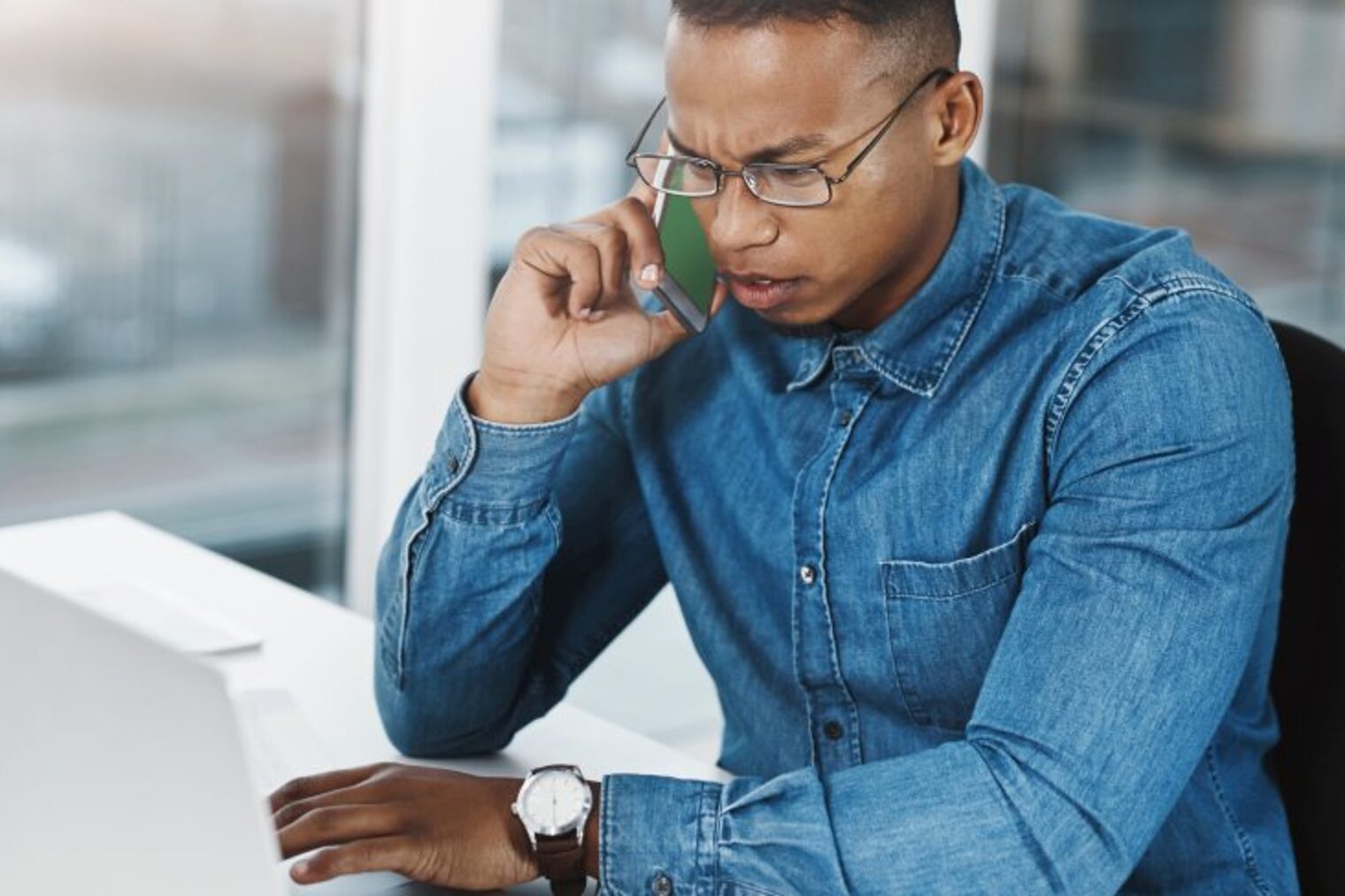 Ein Mann sitzt an einem Schreibtisch vor einem Laptop und telefoniert mit einem Handy. 