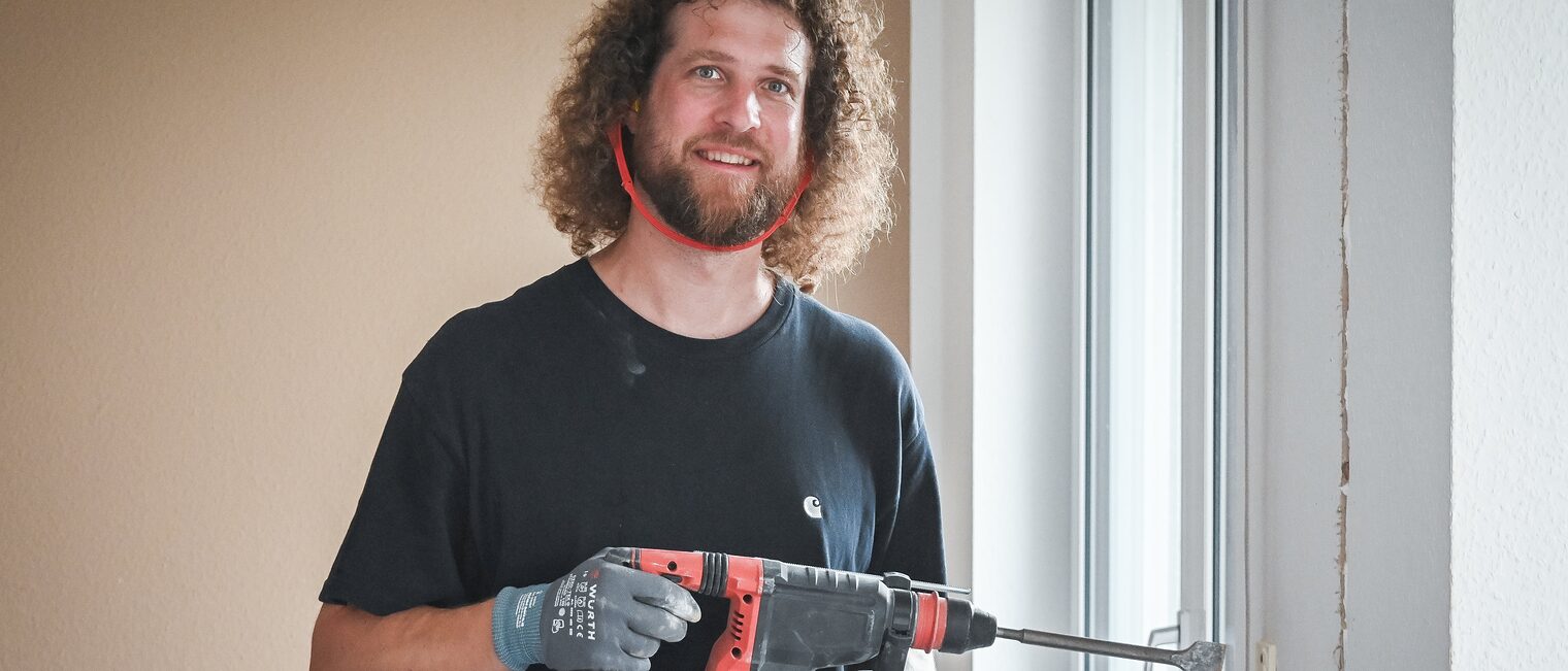 Tobias Haack fühlt sich richtig wohl im Handwerk und nutzt die Möglichkeiten, die es ihm bietet. Ein Mann mit Gehörschutz und einer Bohrmaschine in der Hand lächelt in die Kamera.
