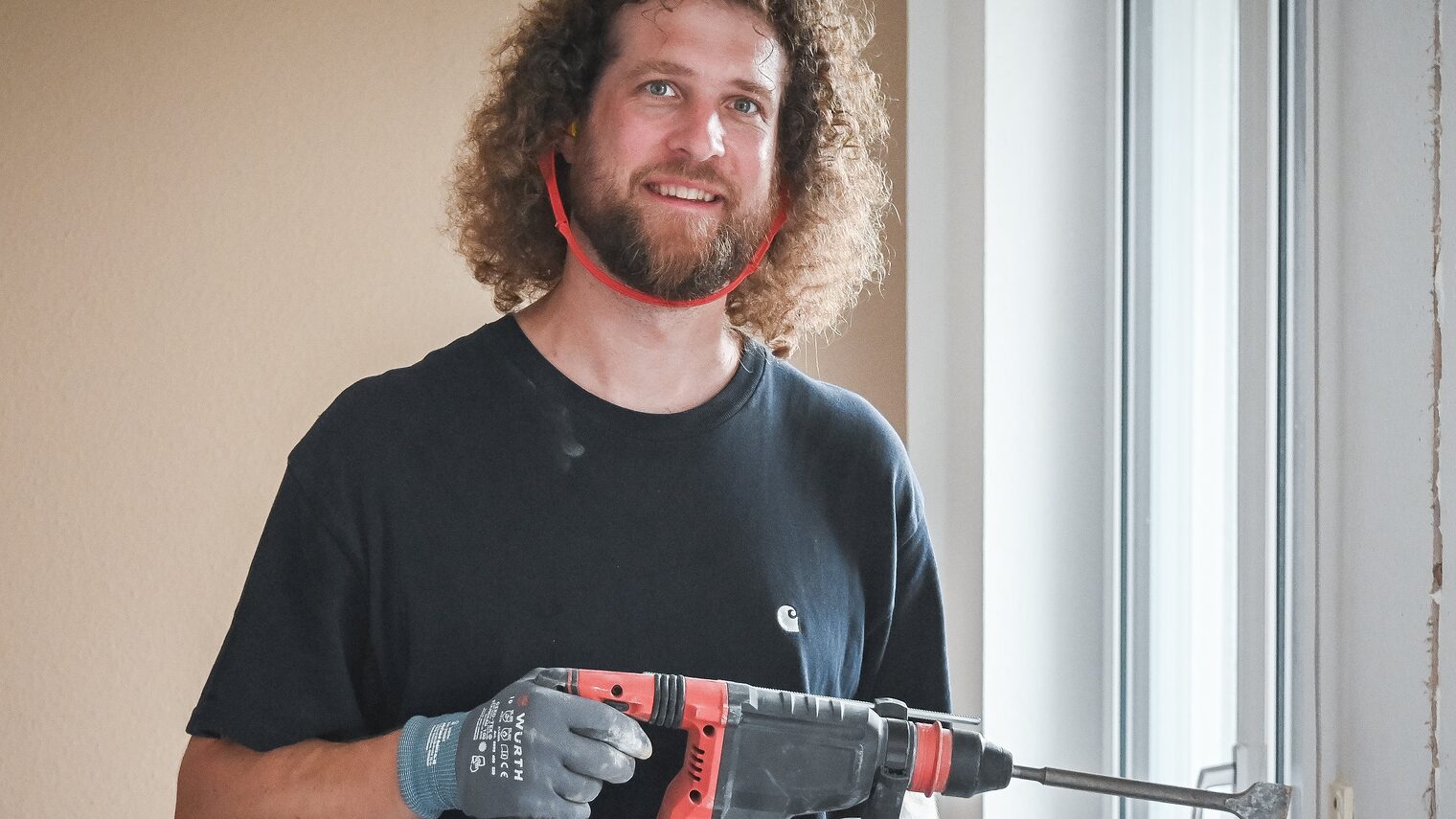 Ein Mann mit Gehörschutz und einer Bohrmaschine in der Hand lächelt in die Kamera. 