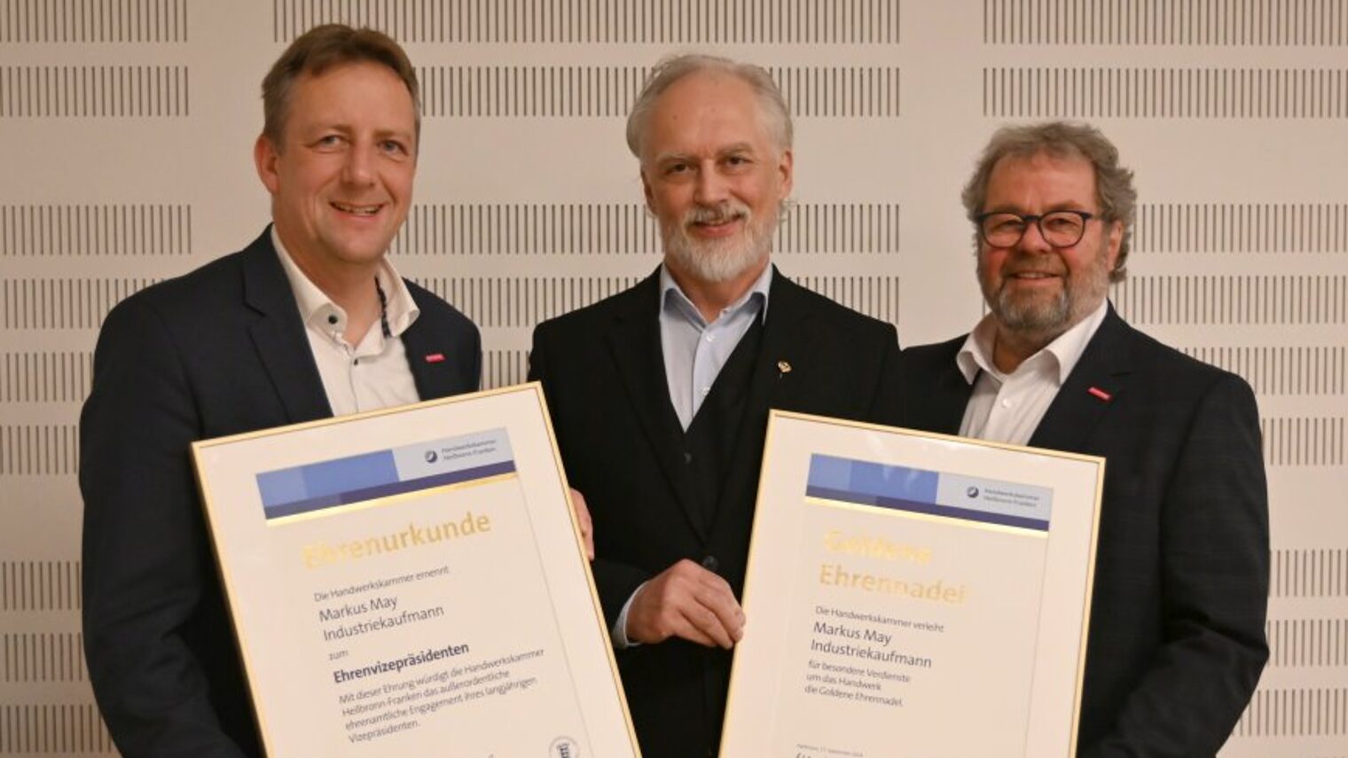 Drei Männer in dunklen Anzügen, zwei Männer halten jeweils eine Urkunde in der Hand.