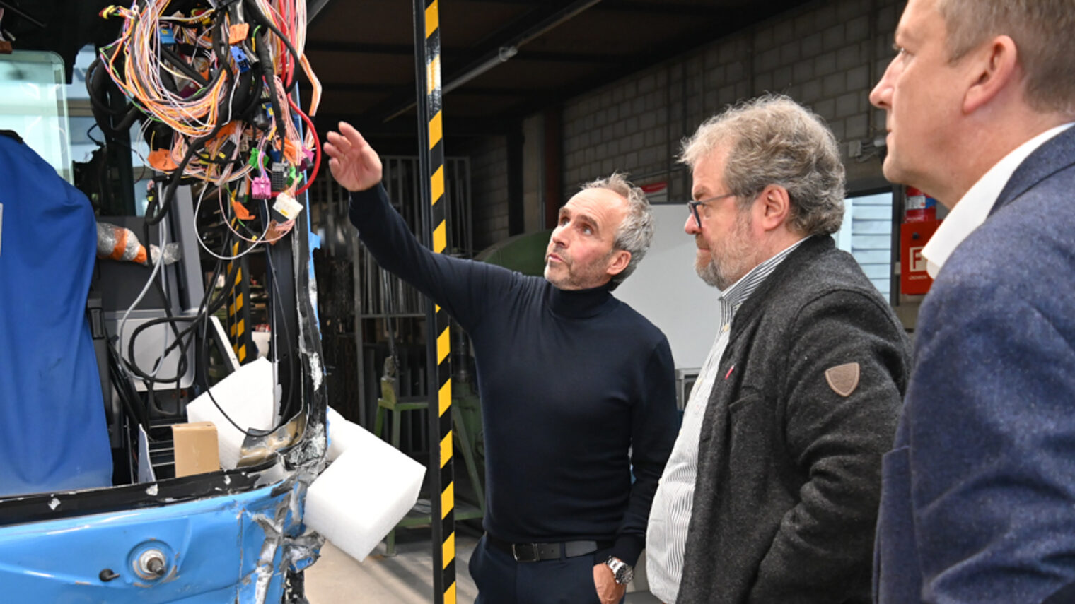 An der Seite steht ein verunfaller Omnibus. Ein Mann zeigt auf den Bus und erklärt zwei Herrn etwas an der Elektronik.