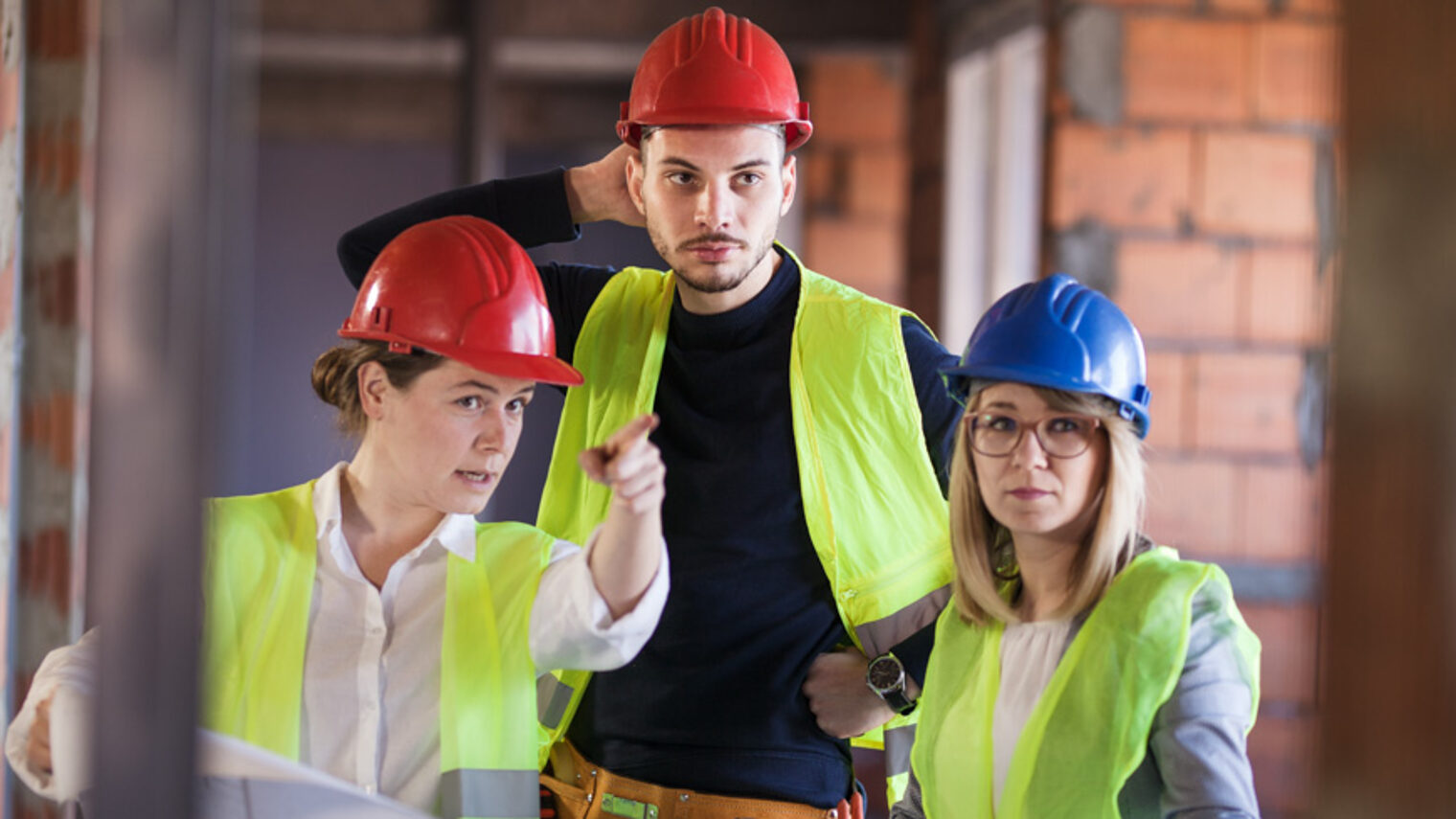 Zwei Frauen und ein Mann stehen in einem Rohbau. Sie tragen Bauhelme und Warnwesten. 