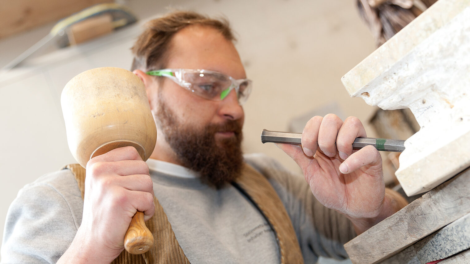 Ein Mann trägt eine Schutzbrille und bearbeitet mit Hammer und Meißel eine Steinplatte.