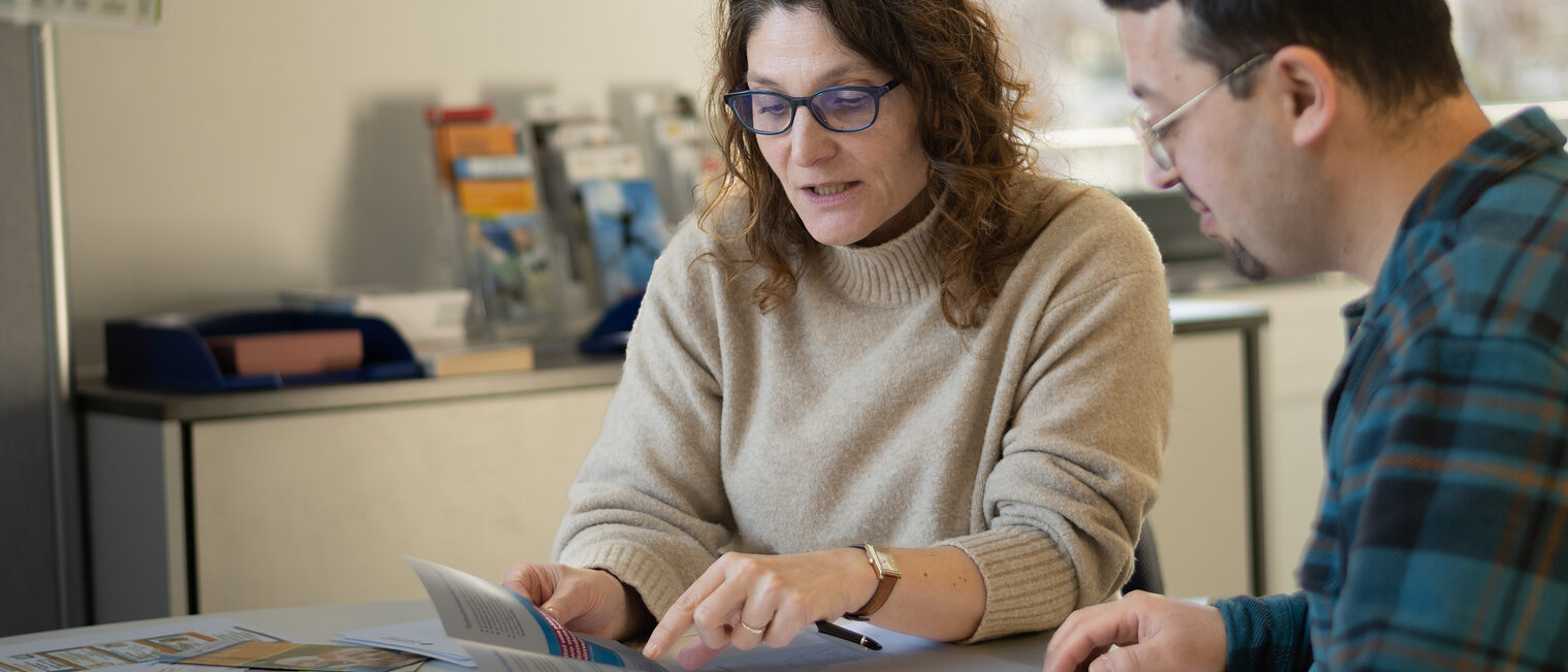 Annabel Gil berät einen Zugewanderten im Rahmen des Kümmerer-Programms. Eine Frau und ein Mann sitzen an einem Tisch. Die Frau erklärt dem Mann etwas anhand eines Programmheftes, auf das sie zeigt.