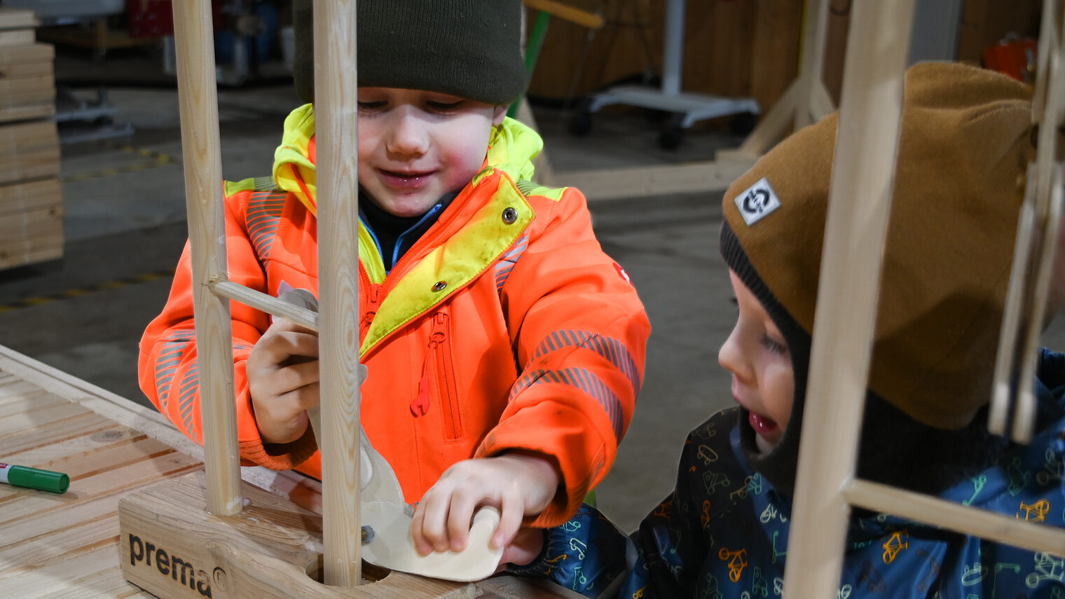 Zwei Kita-Kinder stehen in einer Werkstatt vor einem Holztisch und basteln gemeinsam Kasper-Figuren aus Holz.
