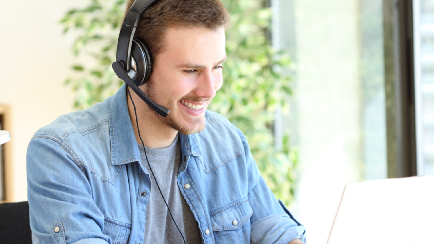 Ein Mann sitzt einem Headset am Laptop und schaut lächelnd in den Monitor