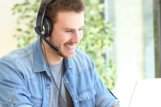 Ein Mann sitzt einem Headset am Laptop und schaut lächelnd in den Monitor