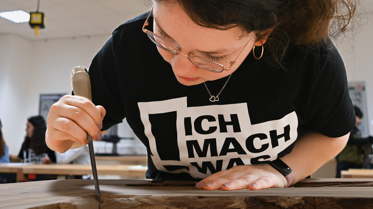 Eine Schülerin steht vor einem Tisch, auf dem ein Werkstück aus Holz liegt, von dem sie mit einem Stemmeisen Leimreste entfernt.