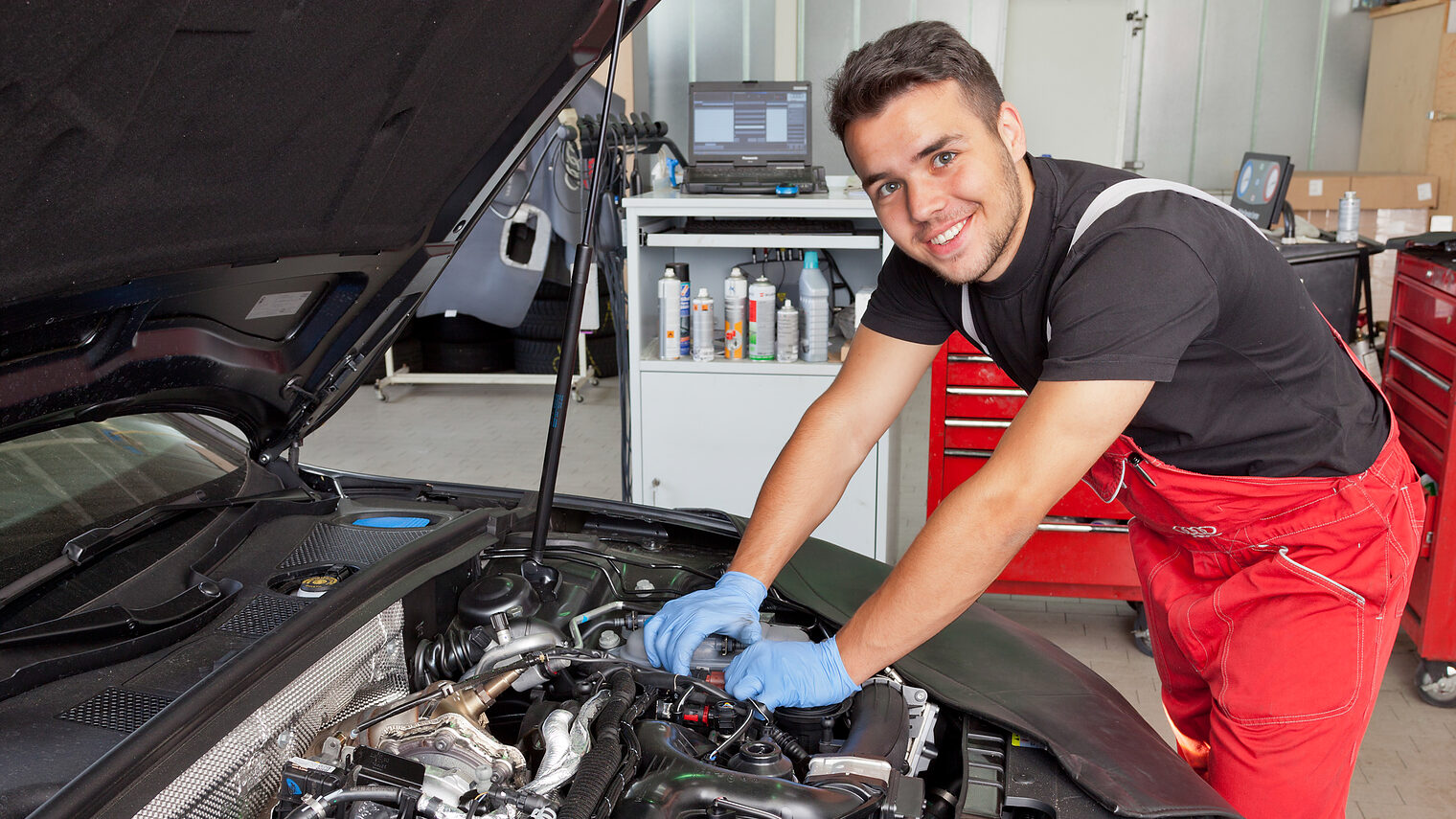 Ein junger Mann, der eine Ausbildung zum Kfz-Mechatroniker absolviert, steht vor einem geöffneten Automotor und repariert ihn.