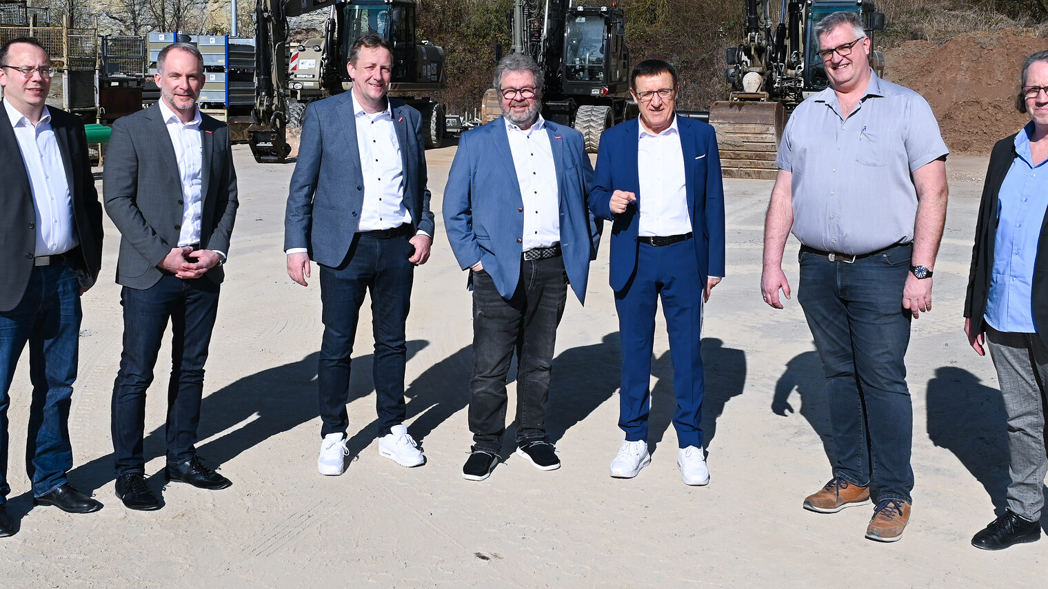 Sieben Männer stehen nebeneinander auf einem Firmengelände und lächeln bei strahlendem Sonnenschein in die Kamera.