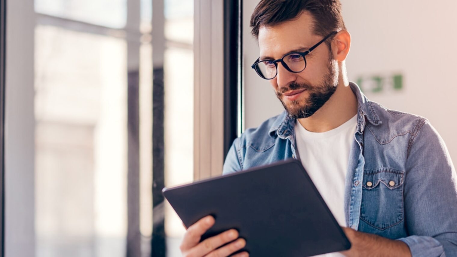 Ein Mann mit einer Brille blickt auf einen Tablet-Bildschirm und steht dabei vor einem großen Fenster.