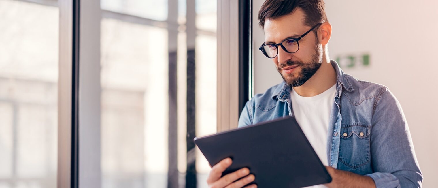 Ein Mann mit einer Brille blickt auf einen Tablet-Bildschirm und steht dabei vor einem großen Fenster.