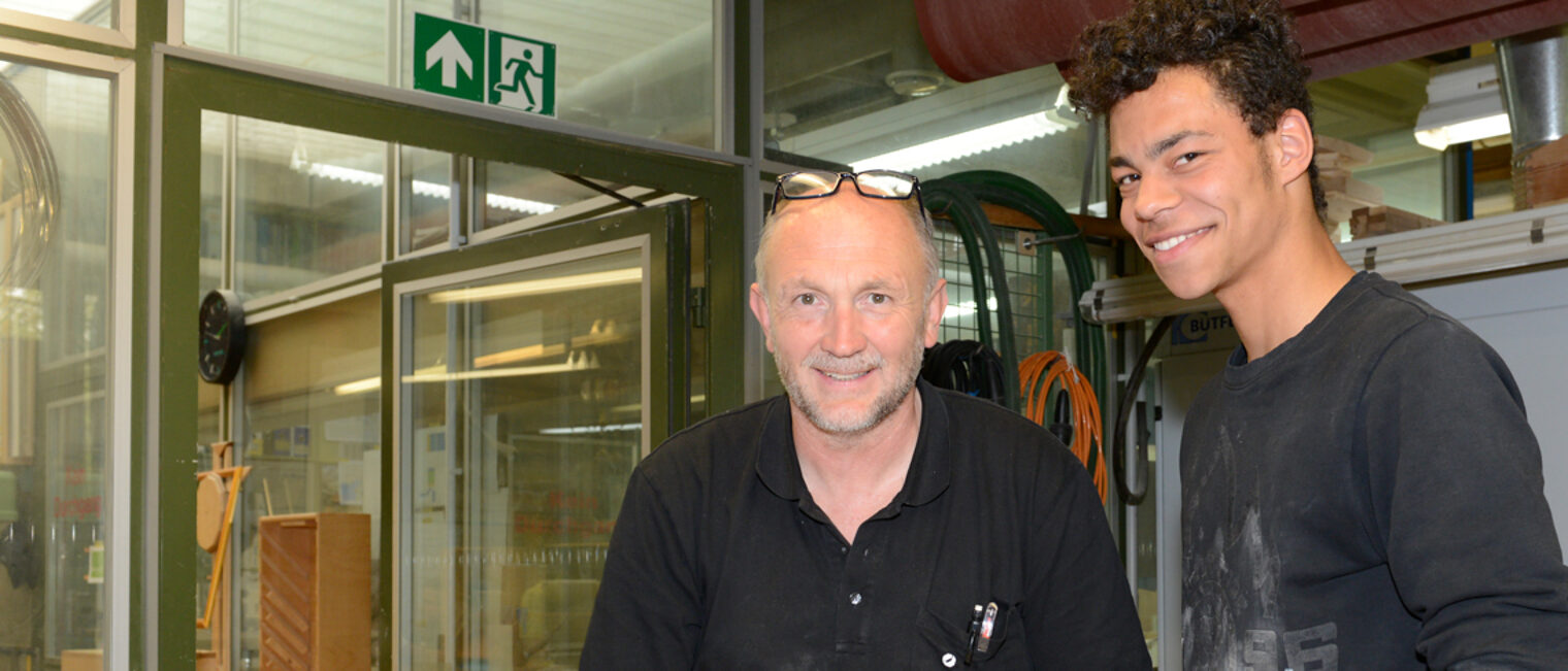 Best Practice gesucht: Integration am Arbeitsmarkt Ausbilder im Handwerk steht zufrieden mit jungem dunkelhäutigem Zugewanderten in der Werkstatt