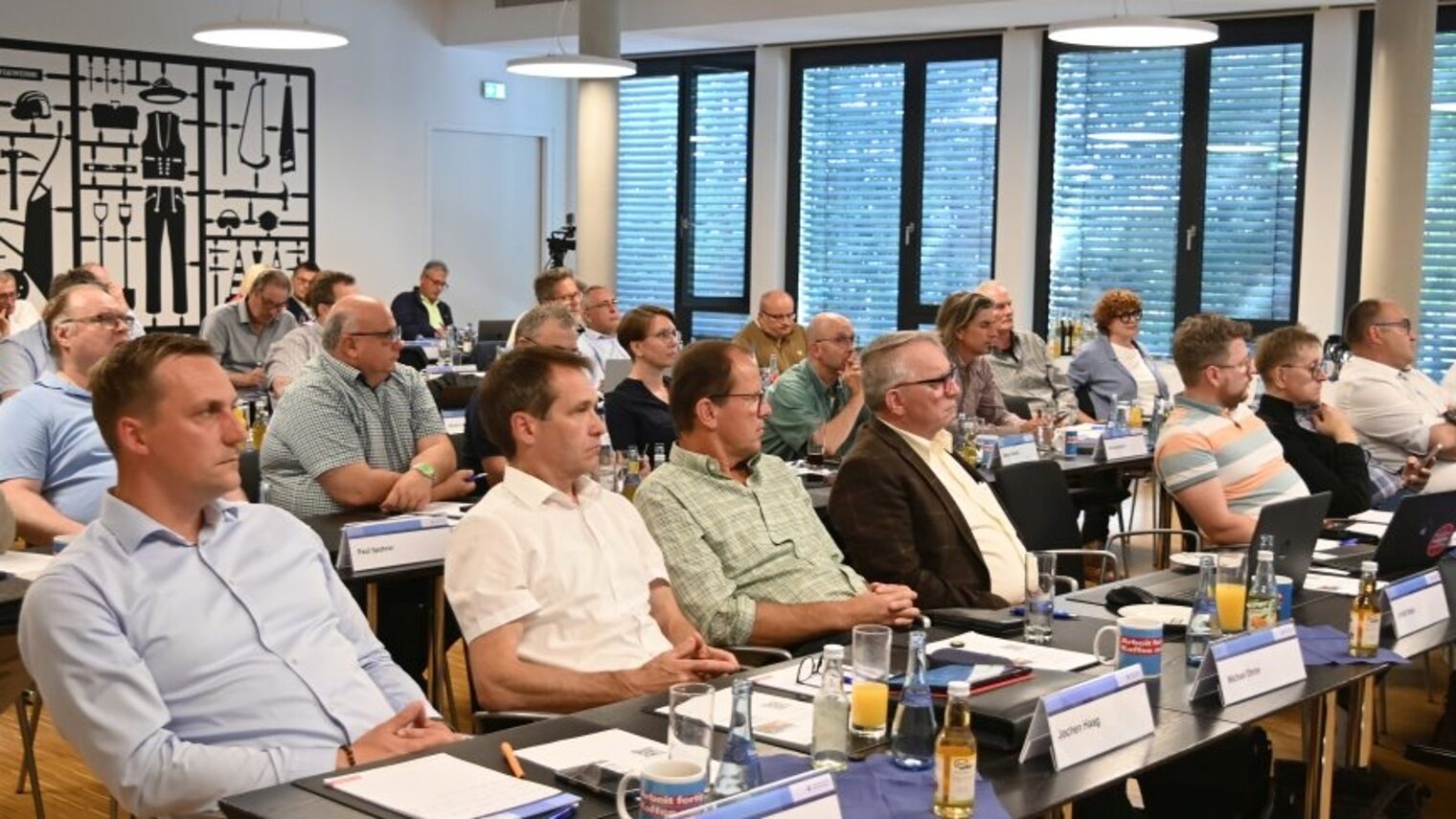 Rund 50 Menschen Sitzen in einem Saal an Tischreihen.
