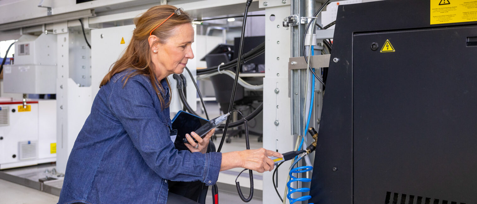 Sabine Worschech, Energieeffizienzmoderatorin von KEFF+, beim Unternehmensrundgang im Rahmen des KEFF+Checks Eine Frau kniet vor einer Maschine und führt eine Druckluftmessung durch.