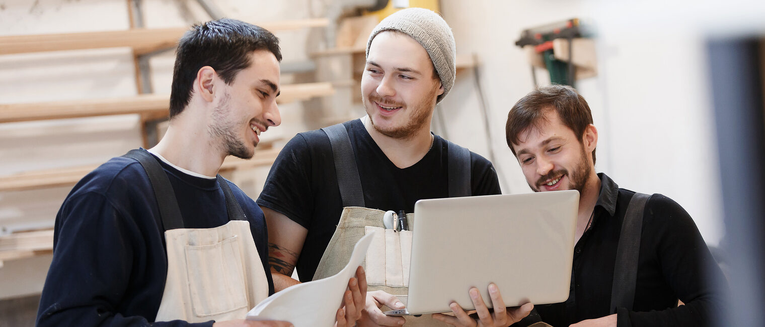 Die Handwerksjunioren wollen dass Handwerk mitgestalten und dafür werben. Drei junge Männer stehen in einer Werkstatt. Der Mann, der in der Mitte steht, hält einen Laptop in den Händen.