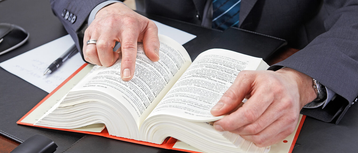 Ein Mann sitzt an einem Schreibtisch. Vor ihm liegt ein aufgeschlagenes Gesetzbuch. Der Mann zeigt auf einen Paragraphen.