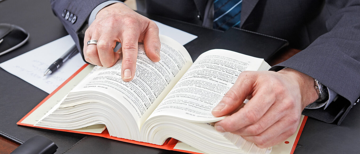 Ein Mann sitzt an einem Schreibtisch. Vor ihm liegt ein aufgeschlagenes Gesetzbuch. Der Mann zeigt auf einen Paragraphen.