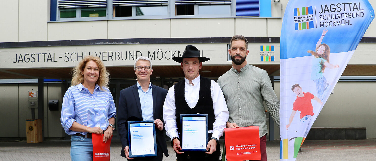 Eine Frau und drei Männer stehen vor einem Schulgebäude. Zwei der Männer halten eine Urkunde in den Händen.